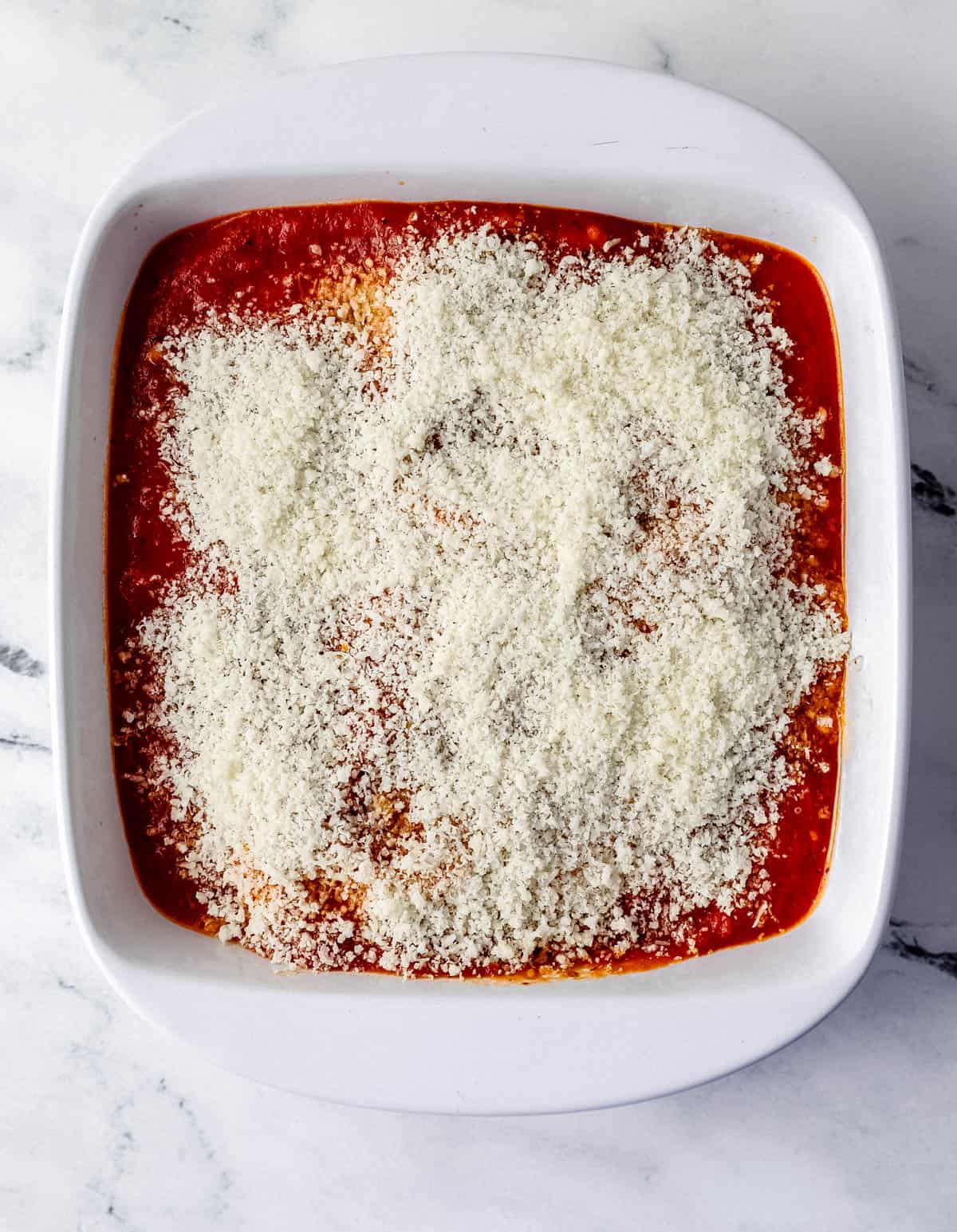 Chicken and sauce topped with grated parmesan in white baking dish on marble surface. 