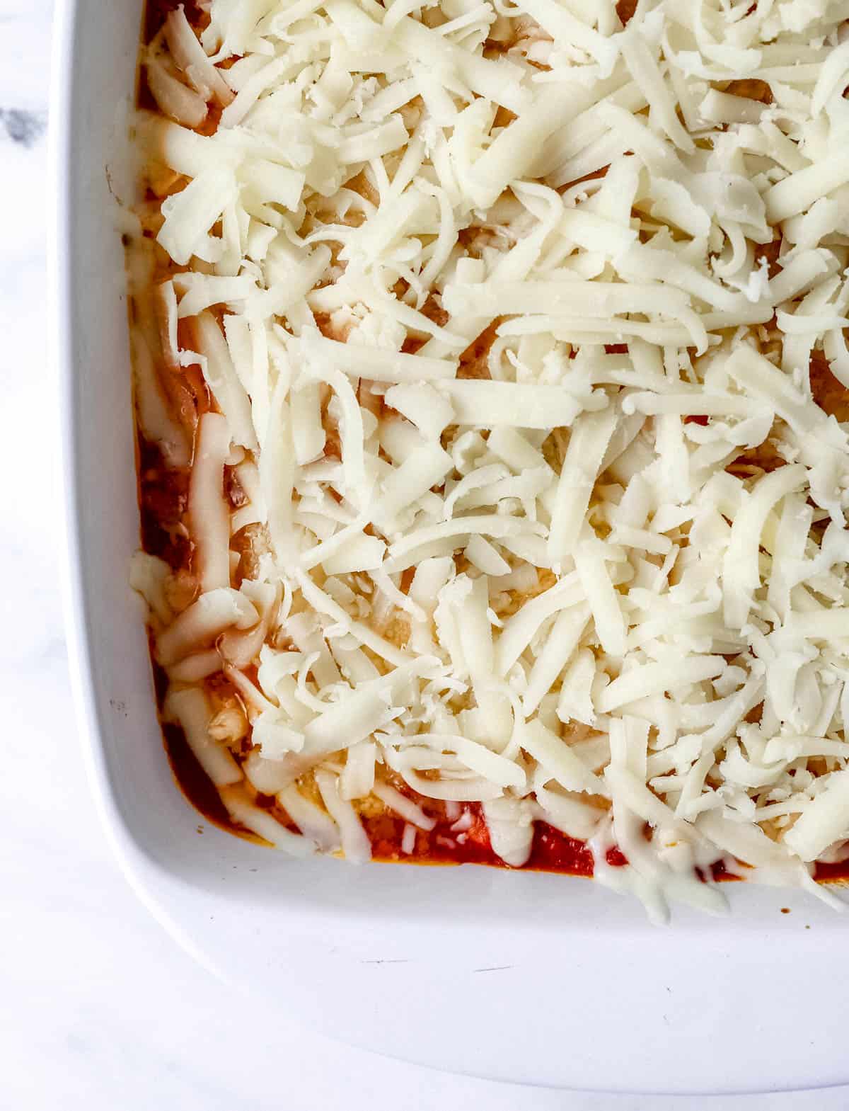 Close up of shredded mozzarella cheese added to chicken, sauce, and parmesan in white baking dish. 