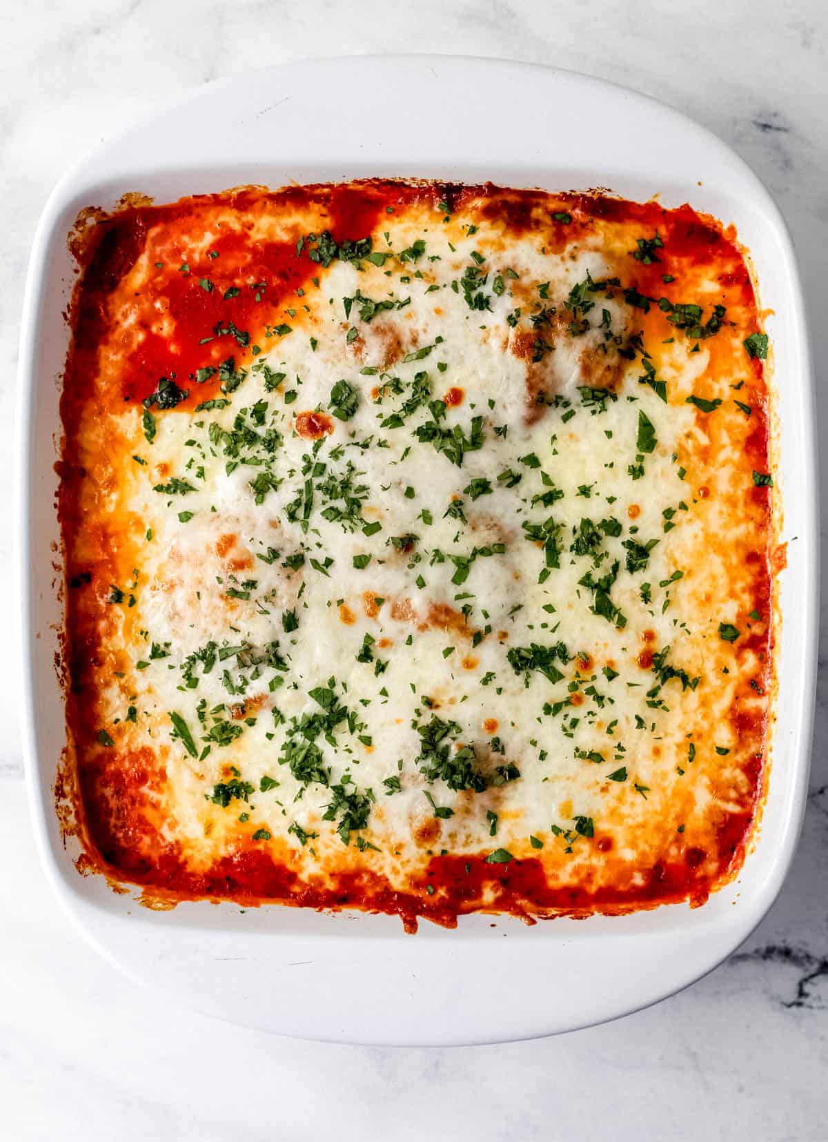 Overhead view of chicken mozzarella bake in square white baking dish on marble surface topped with chopped parsley. 