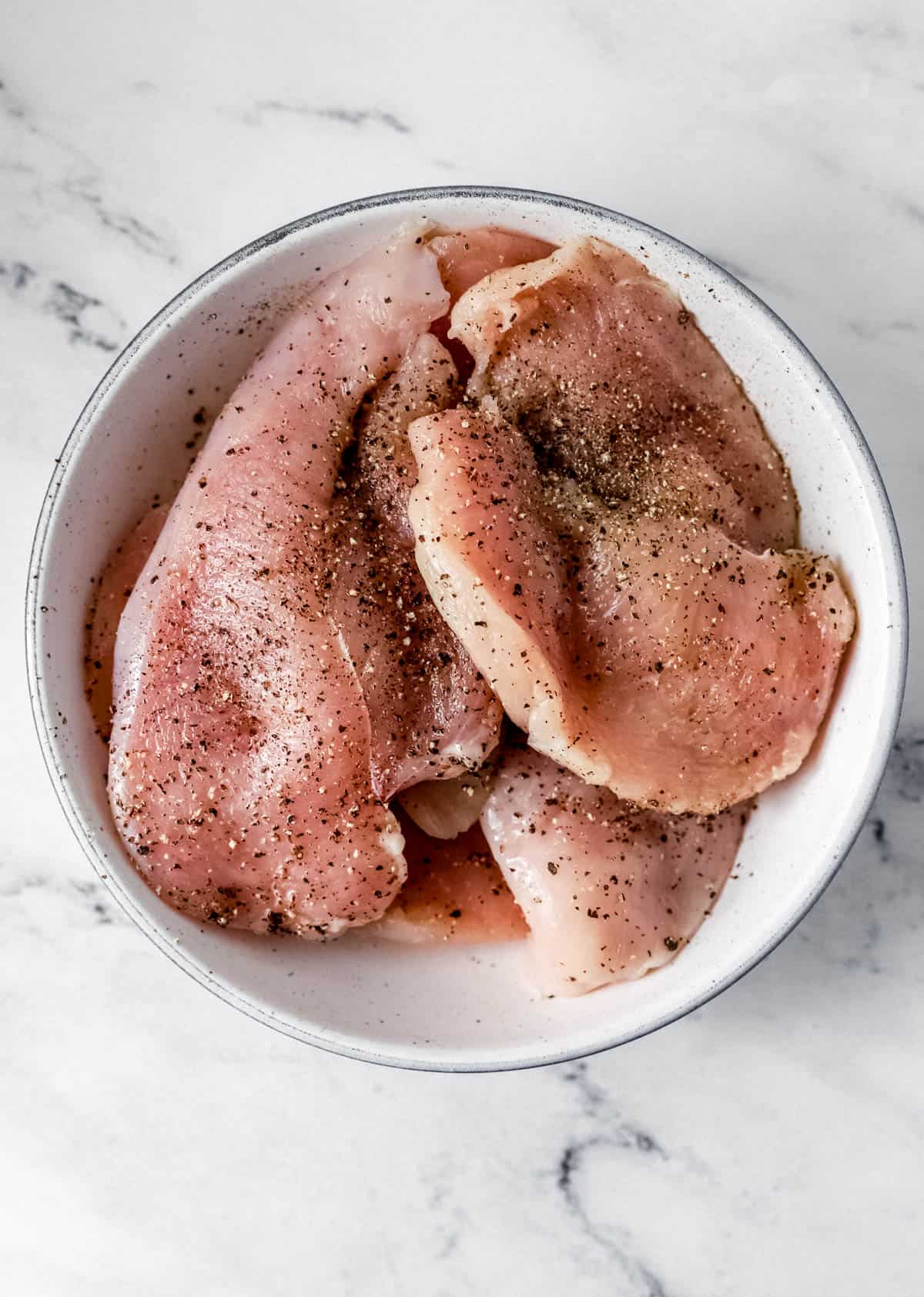 Raw chicken seasoned with salt and ground black pepper in white bowl on marble surface. 