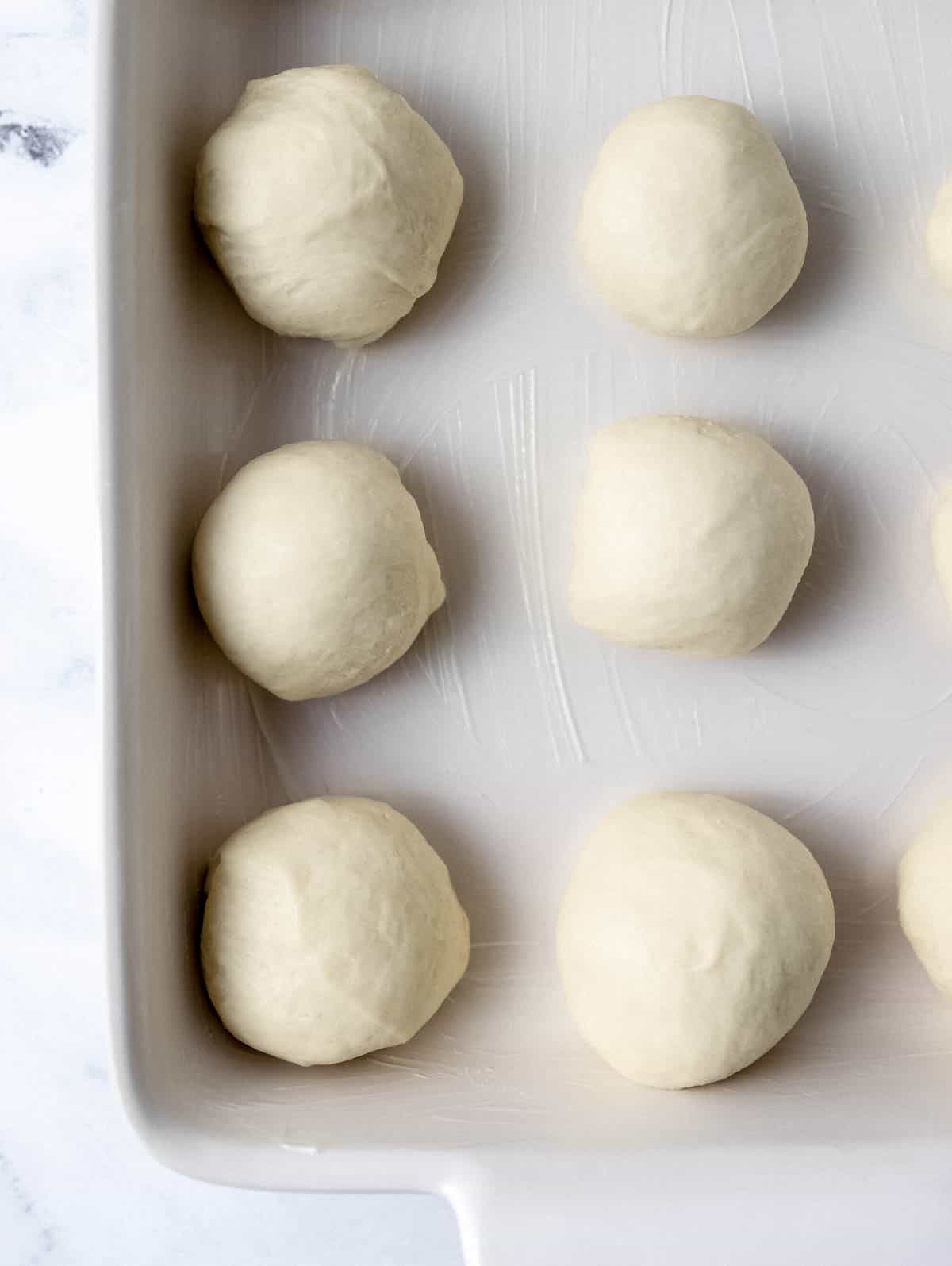 Dough balls added to buttered baking dish on marble surface. 
