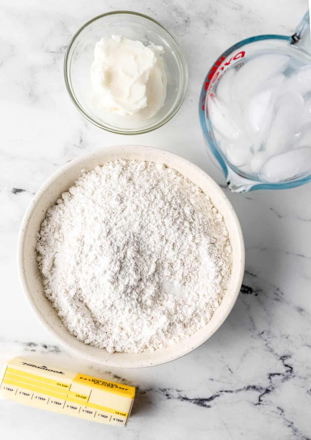 Ingredients needed to make pie crust in separate containers on marble surface. 