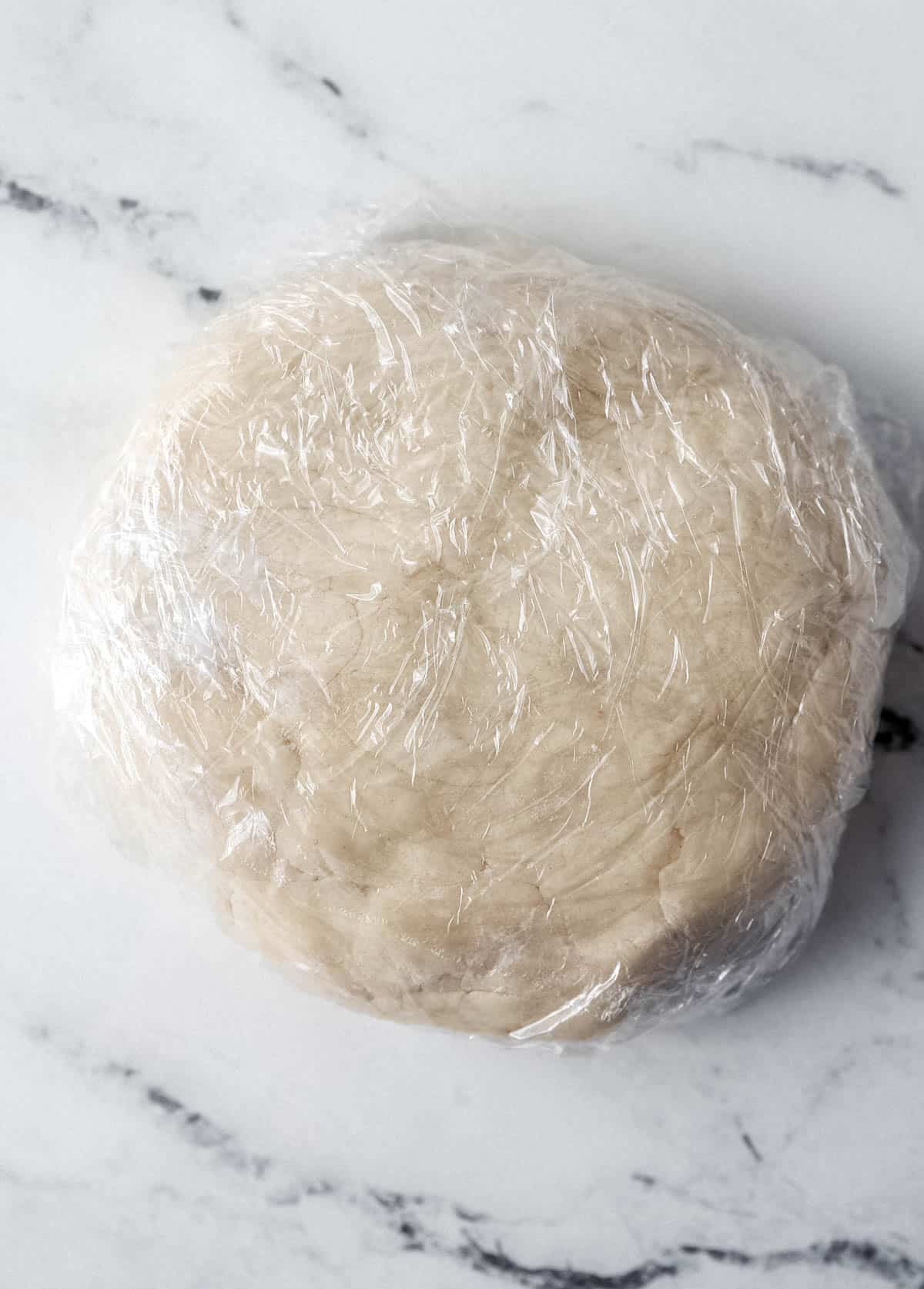 Pie dough wrapped in plastic wrap on marble surface. 