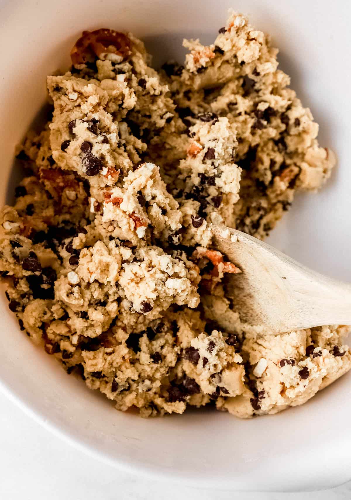 Chocolate chips and pretzels added to cookie dough in white bowl. 