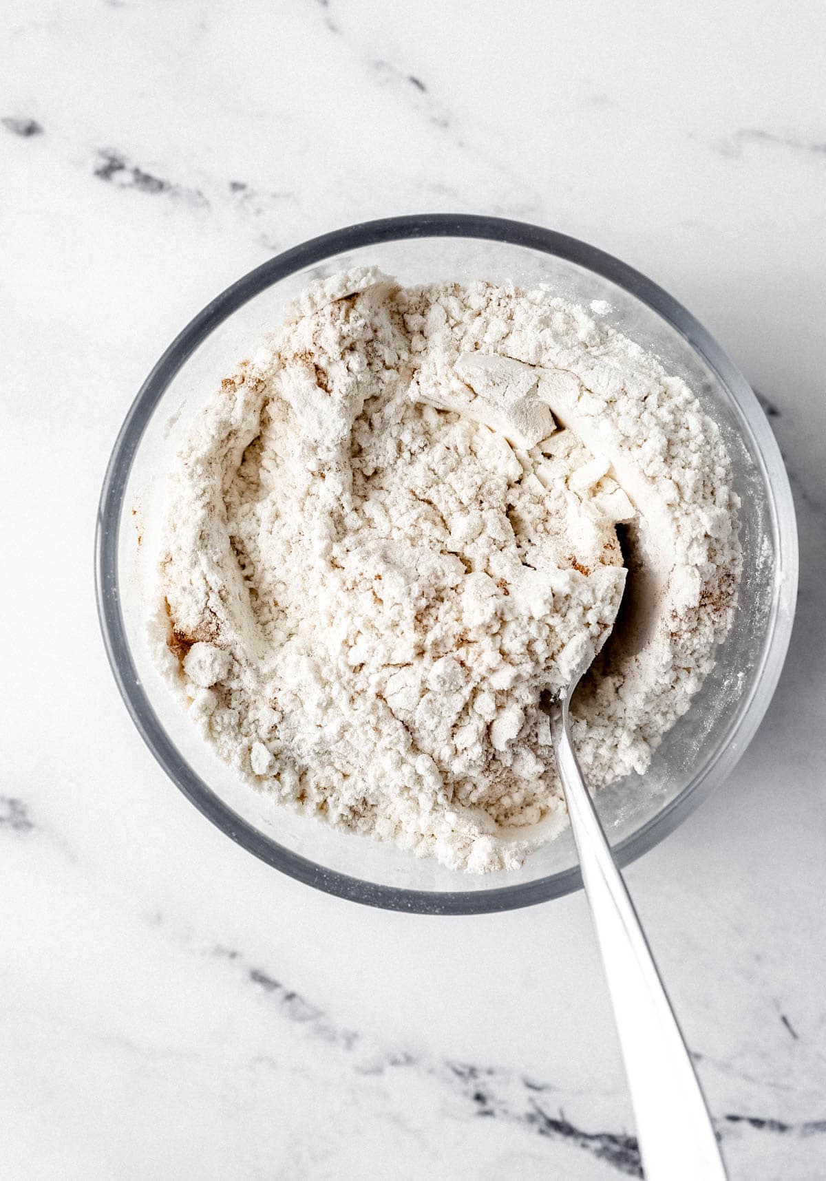 Flour mixture to make muffins in glass bowl with spoon on marble surface.