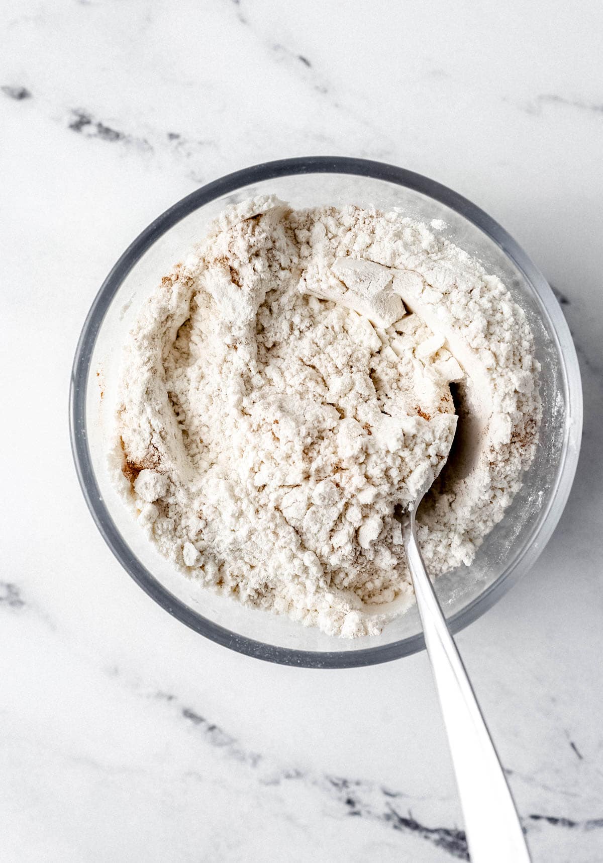 Flour mixture to make muffins in glass bowl with spoon on marble surface. 