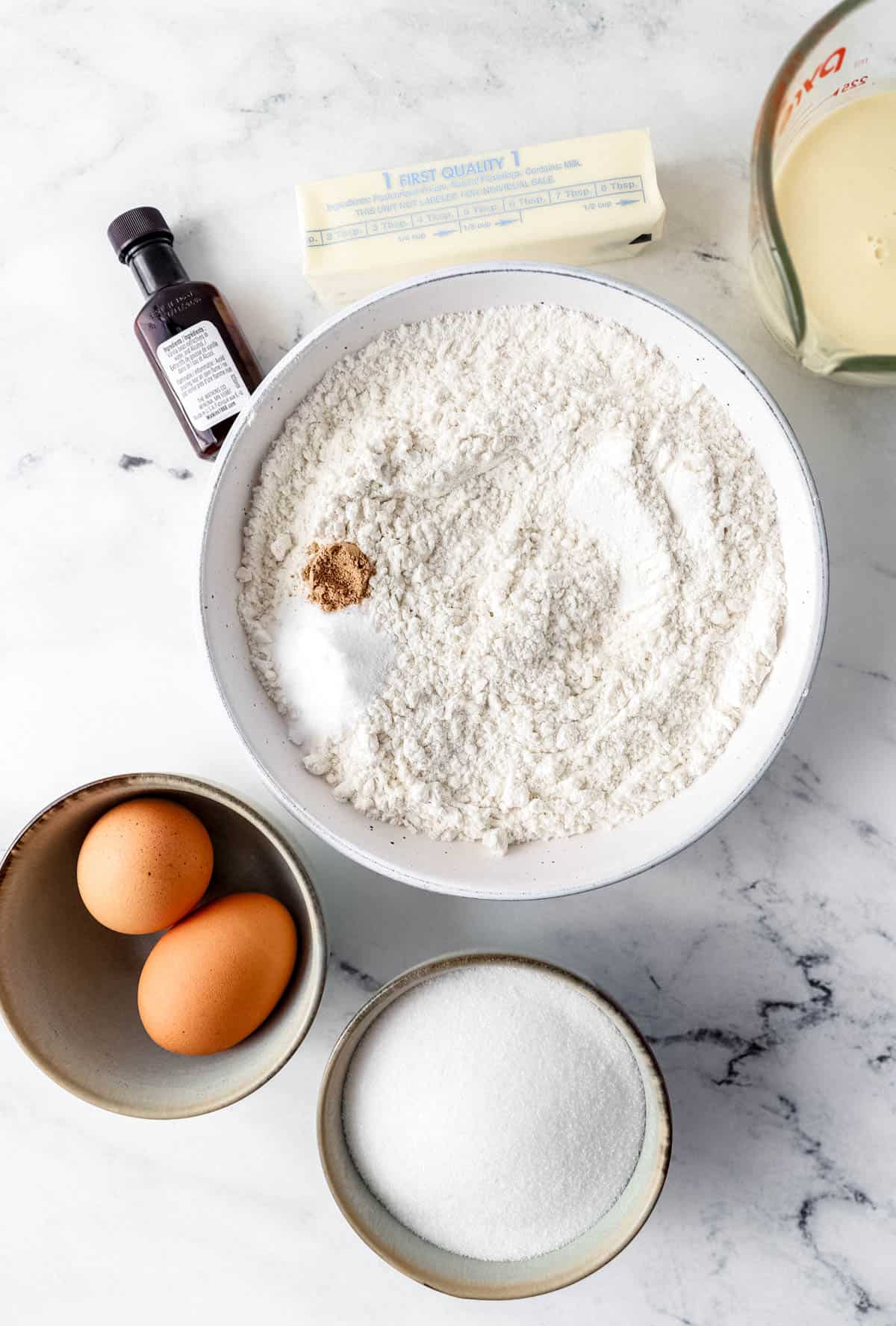 Overhead view of ingredients needed to make eggnog cake in separate bowls and containers on marble surface. 