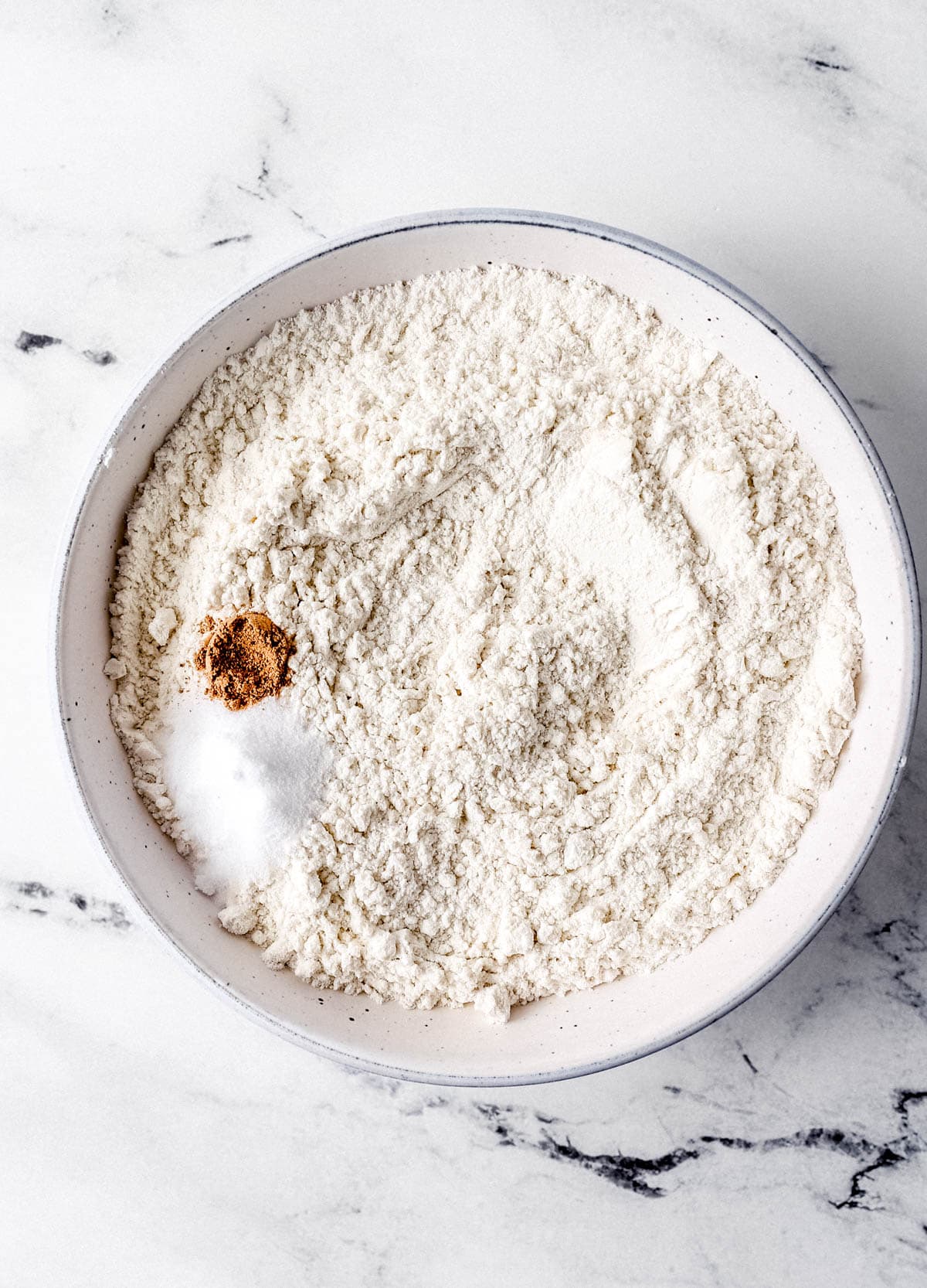 Overhead view of dry ingredients to make cake in white bowl on marble surface. 