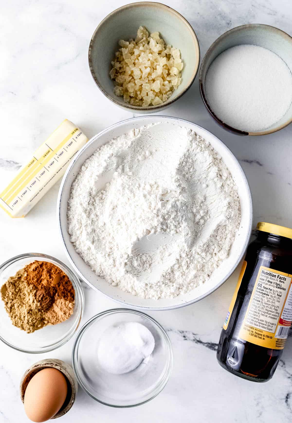 Overhead view of ingredients needed to make gingerbread cake in separate bowls and containers on marble surface. 