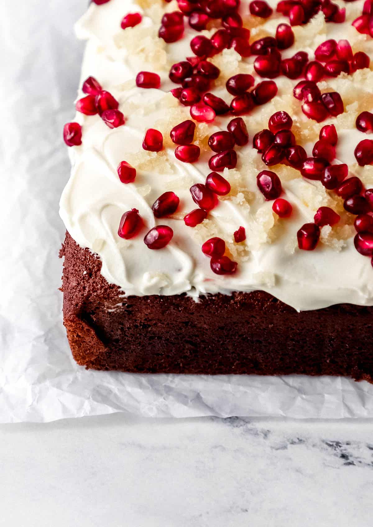 Close up view of finished gingerbread cake on parchment paper topped with frosting, pomegranate arils, and chopped candied ginger. 