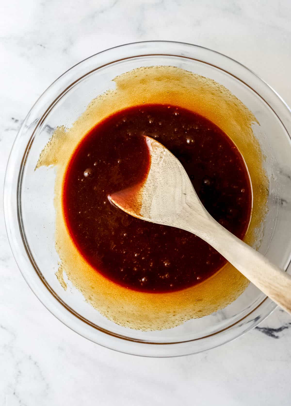 Overhead view of wet cake ingredients combined in glass mixing bowl with wooden spoon. 