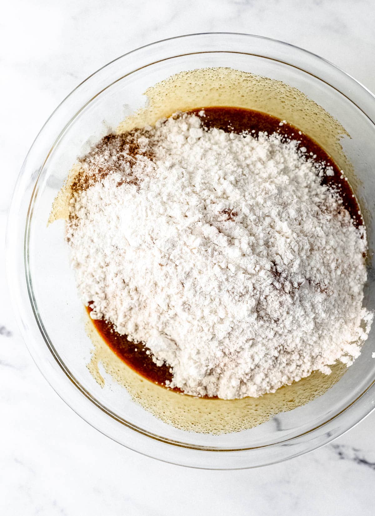 Overhead view of flour mixture added to wet ingredients in large glass mixing bowl. 