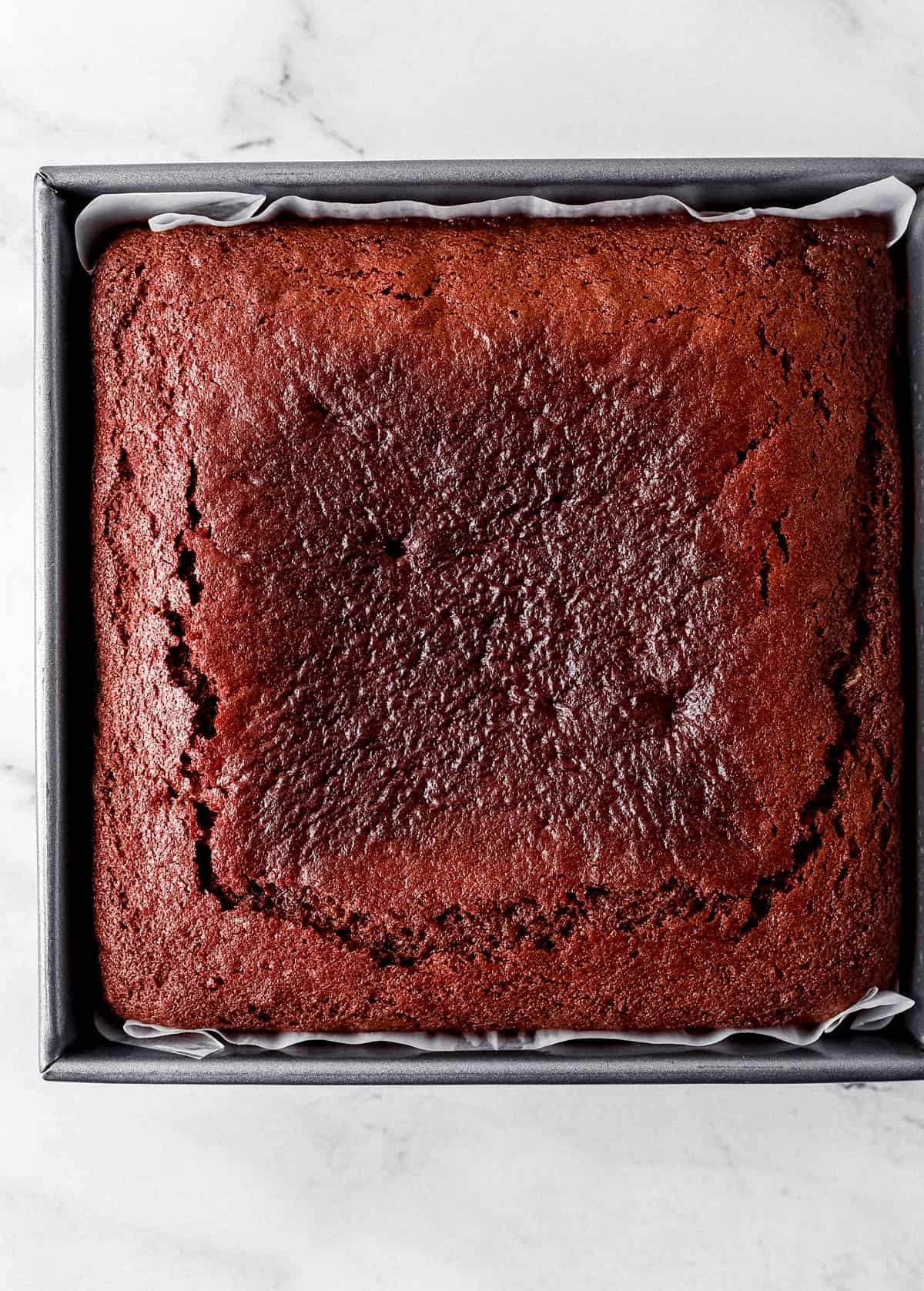 Overhead view of baked gingerbread cake in parchment lined baking pan on marble surface. 