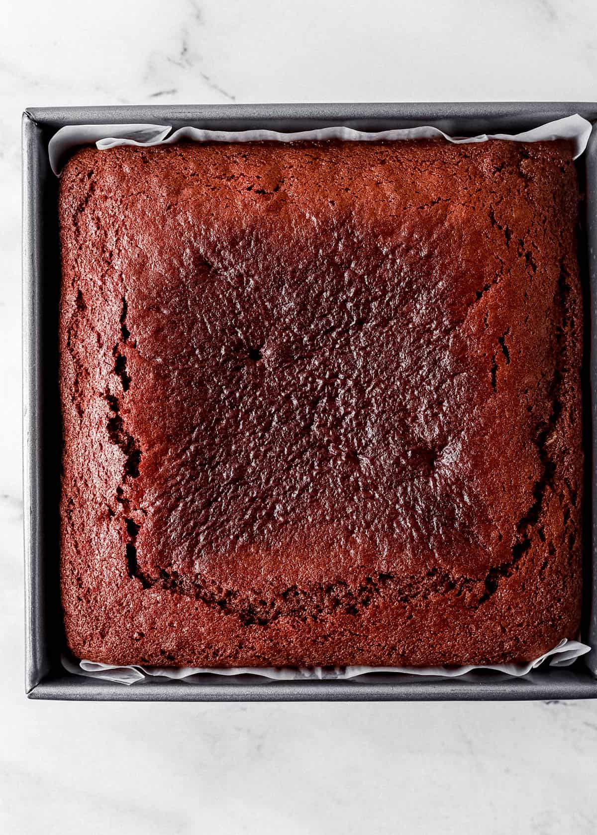 Overhead view of baked gingerbread cake in parchment lined baking pan on marble surface.