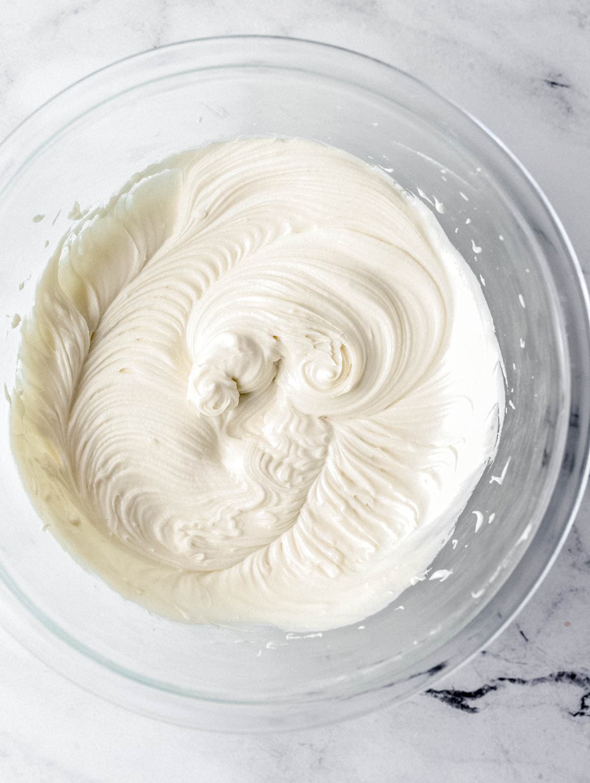 Cake frosting ingredients combined in large glass mixing bowl on marble surface.