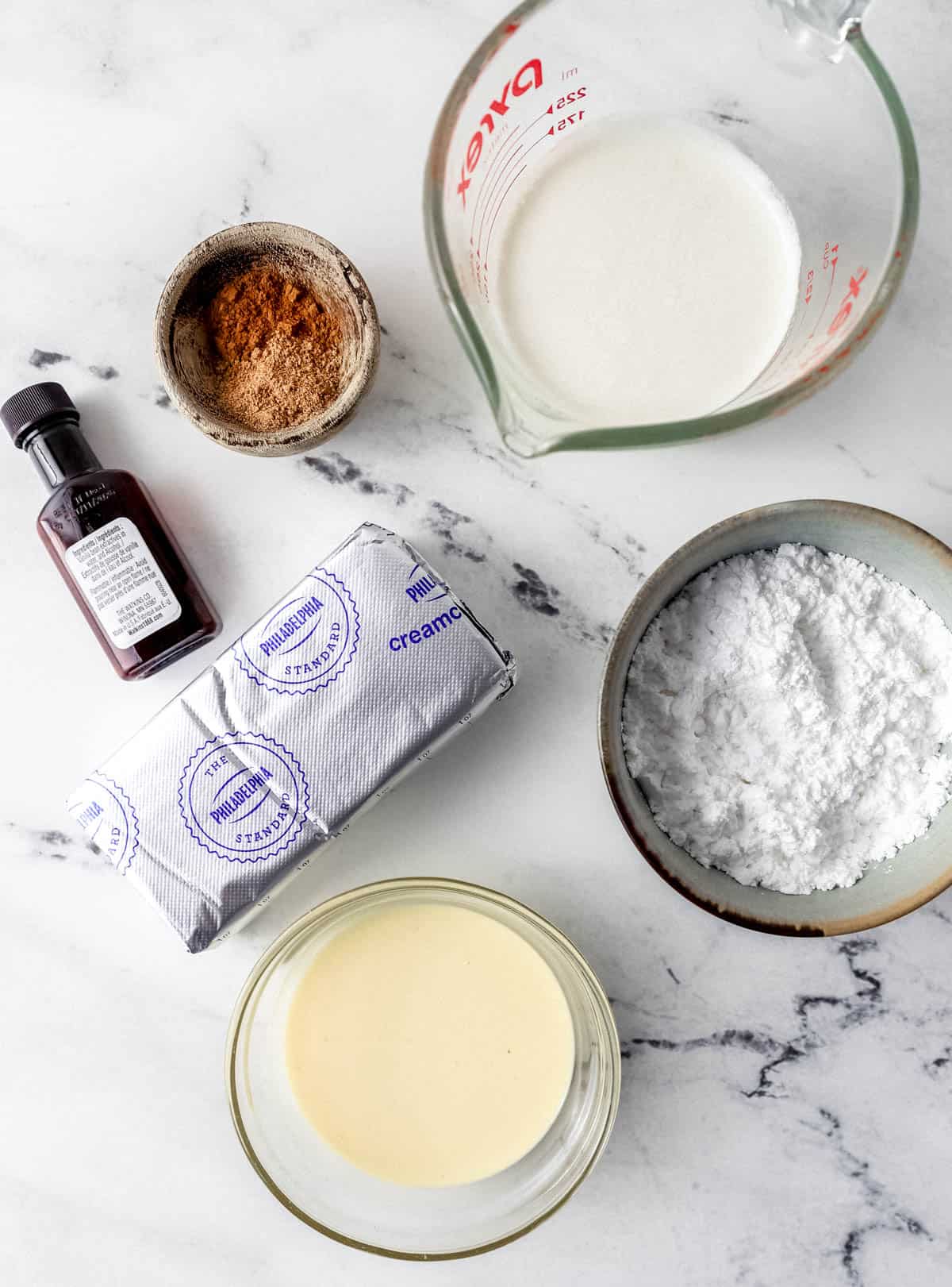 Overhead view of ingredients needed to make cheesecake in separate containers on marble surface. 