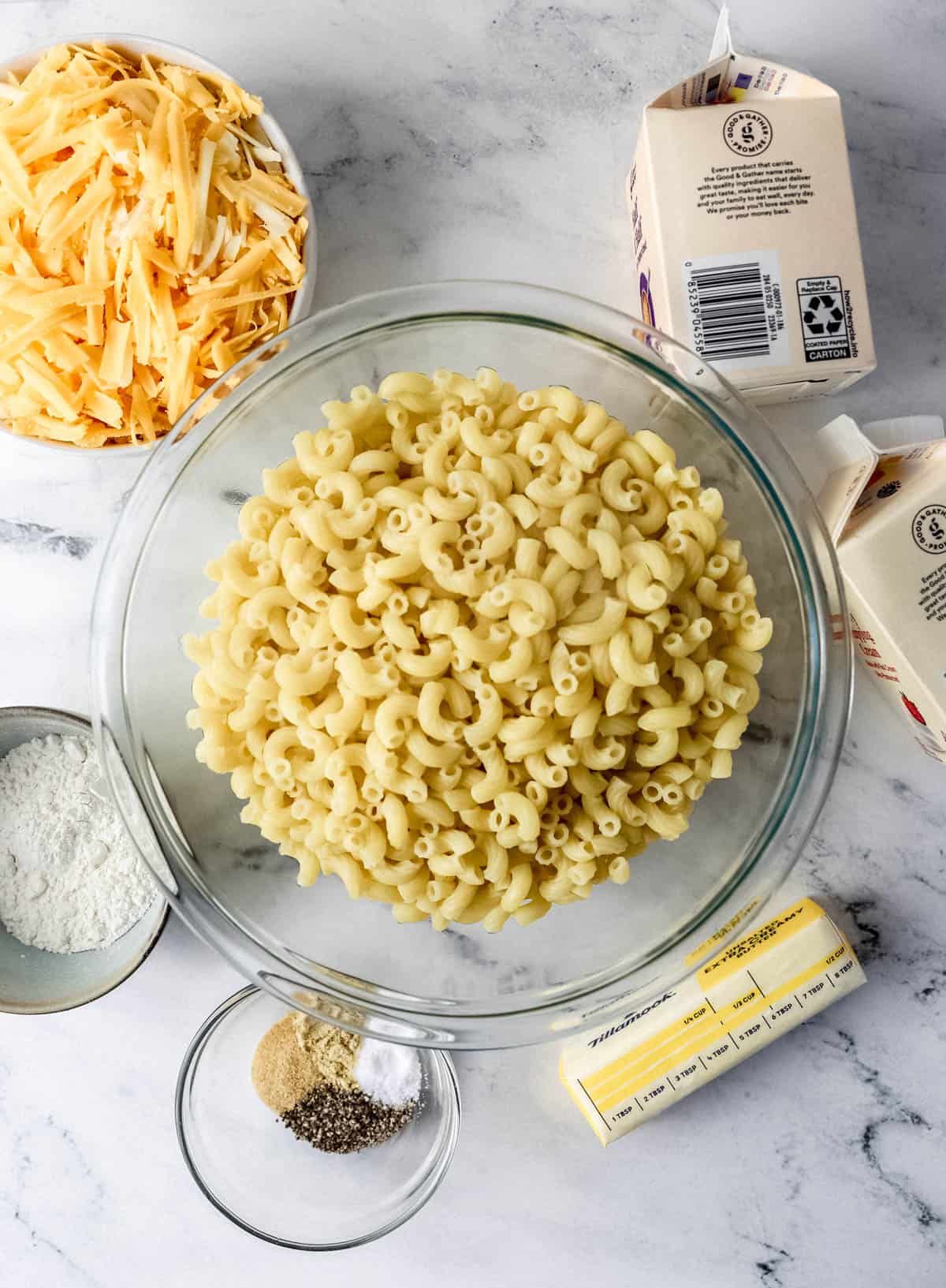 Overhead view of ingredients needed to make mac and cheese in separate containers on marble surface. 