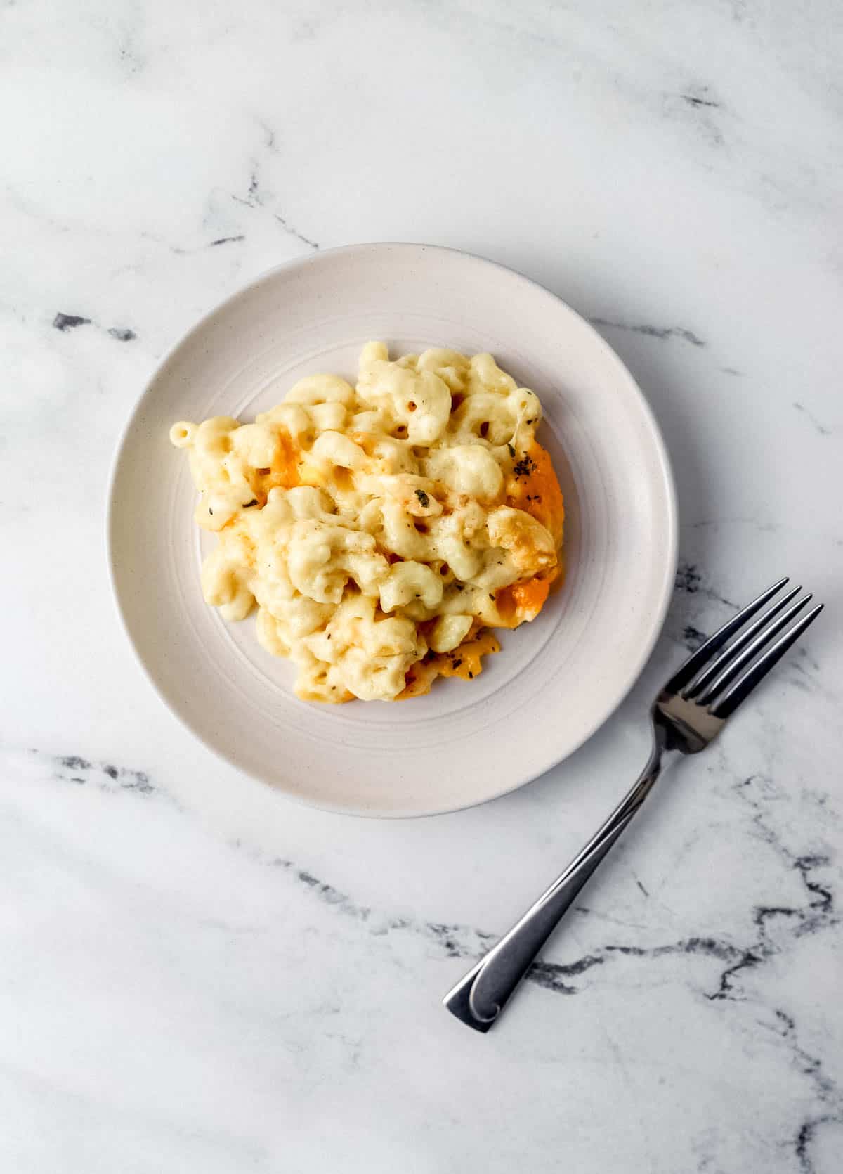 Overhead view of white plate with serving of mac and cheese on it beside a for on marble surface. 