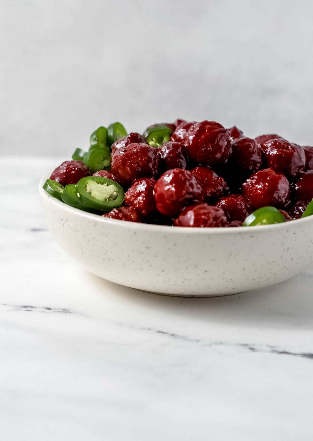 Close up side view of finished meatballs and jalapeno slices in white bowl on marble surface. 