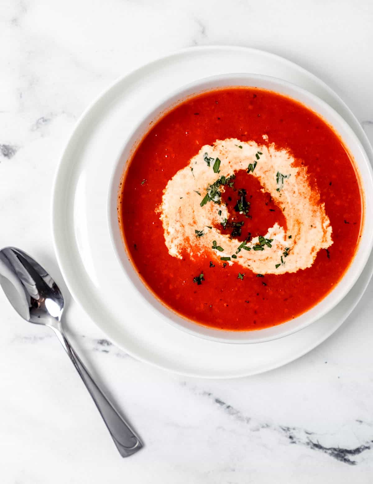 White bowl of soup on white plate beside a spoon on marble surface. 
