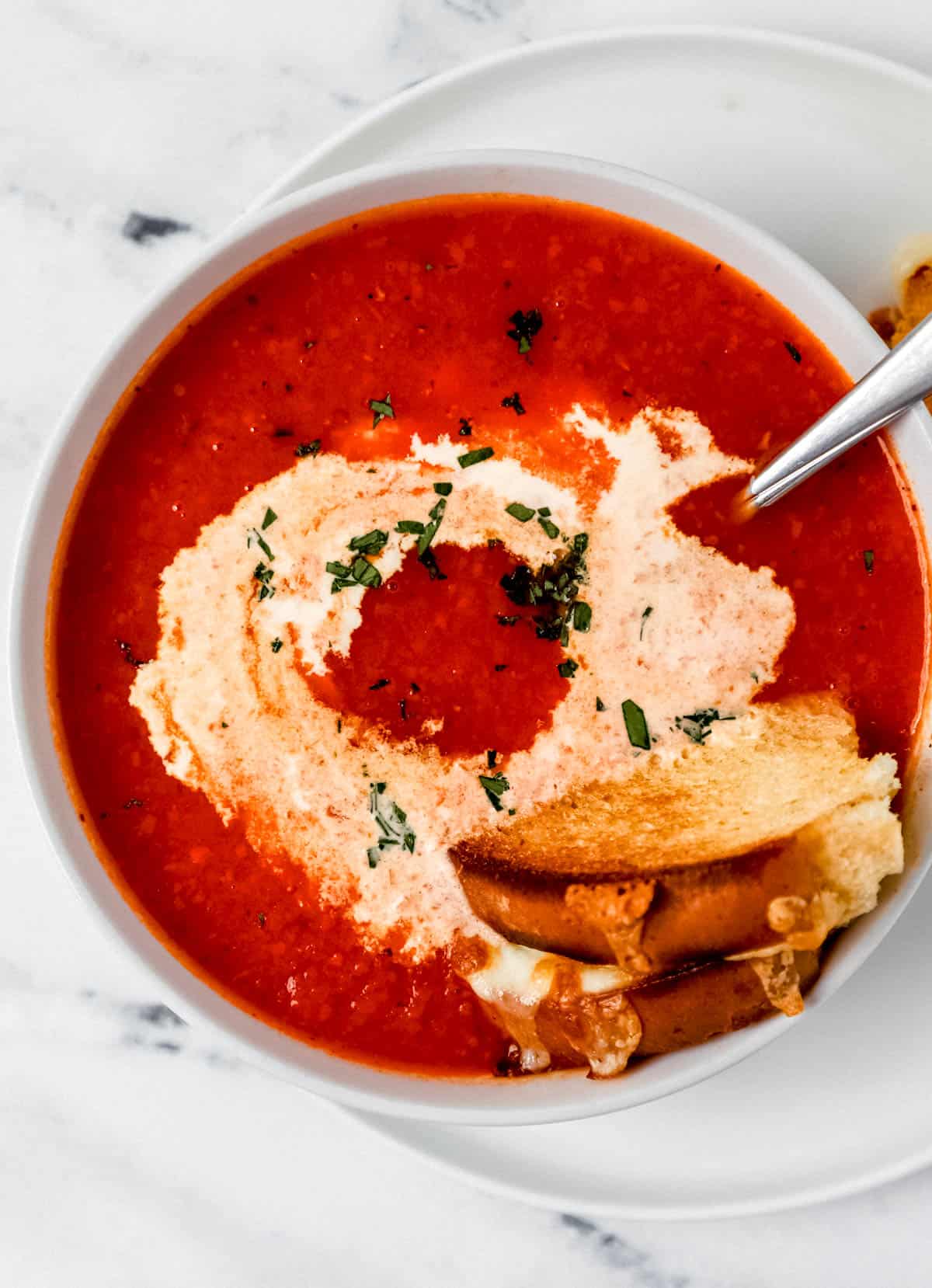 Overhead view of white bowl of tomato soup on white plate with a spoon in it topped with cream and a grilled cheese.