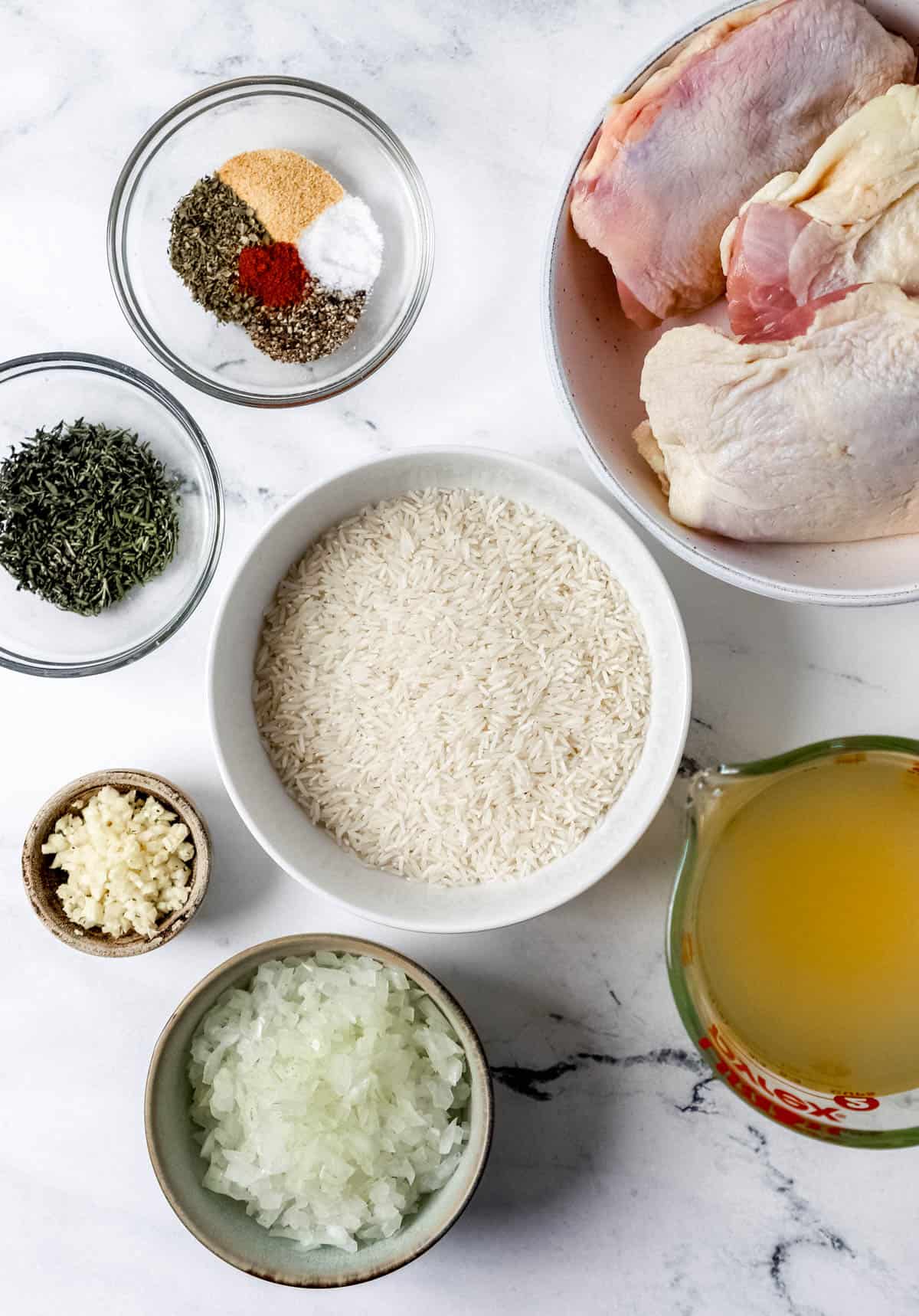 Overhead view of ingredients needed to make chicken and rice in separate bowls on marble surface. 
