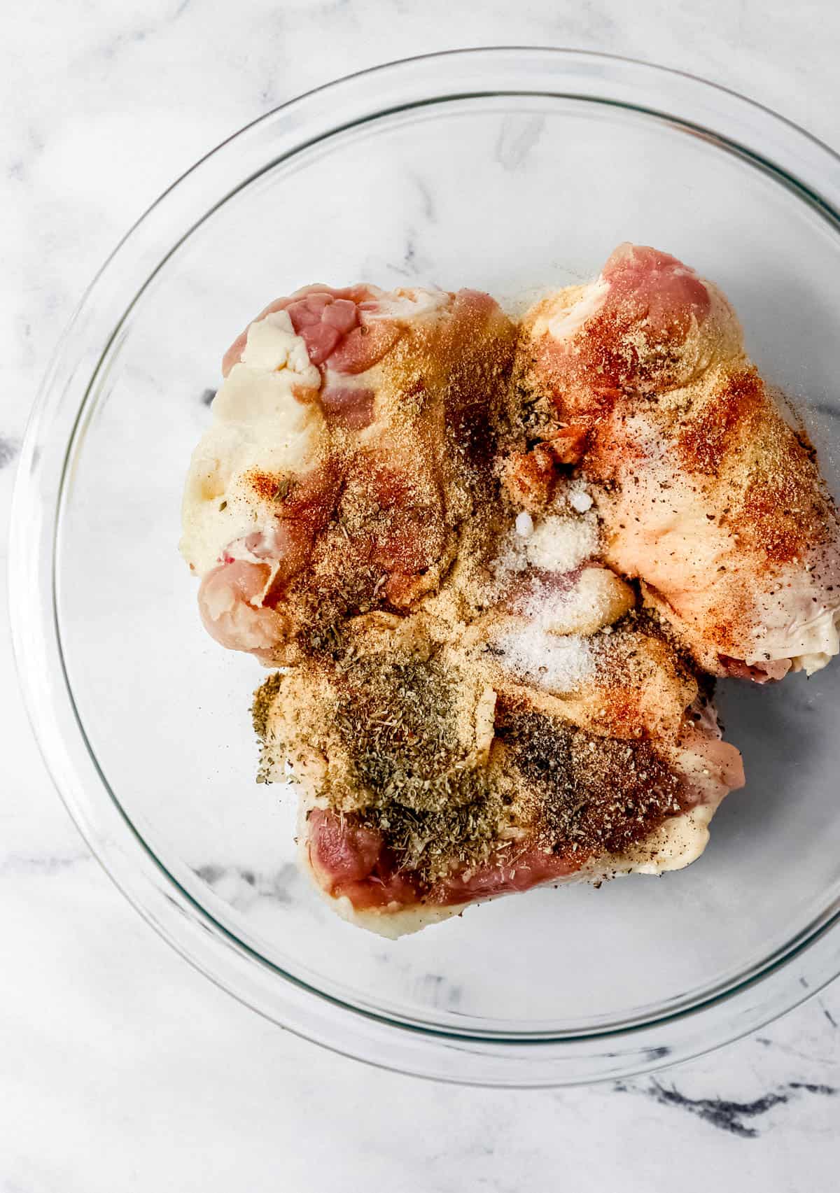 Overhead view of raw chicken with seasoning in glass mixing bowl. 