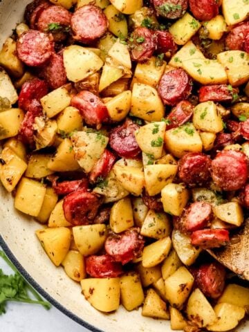 Overhead view of finished sausage and potatoes recipe in white braiser on marble surface with wooden spoon in it.