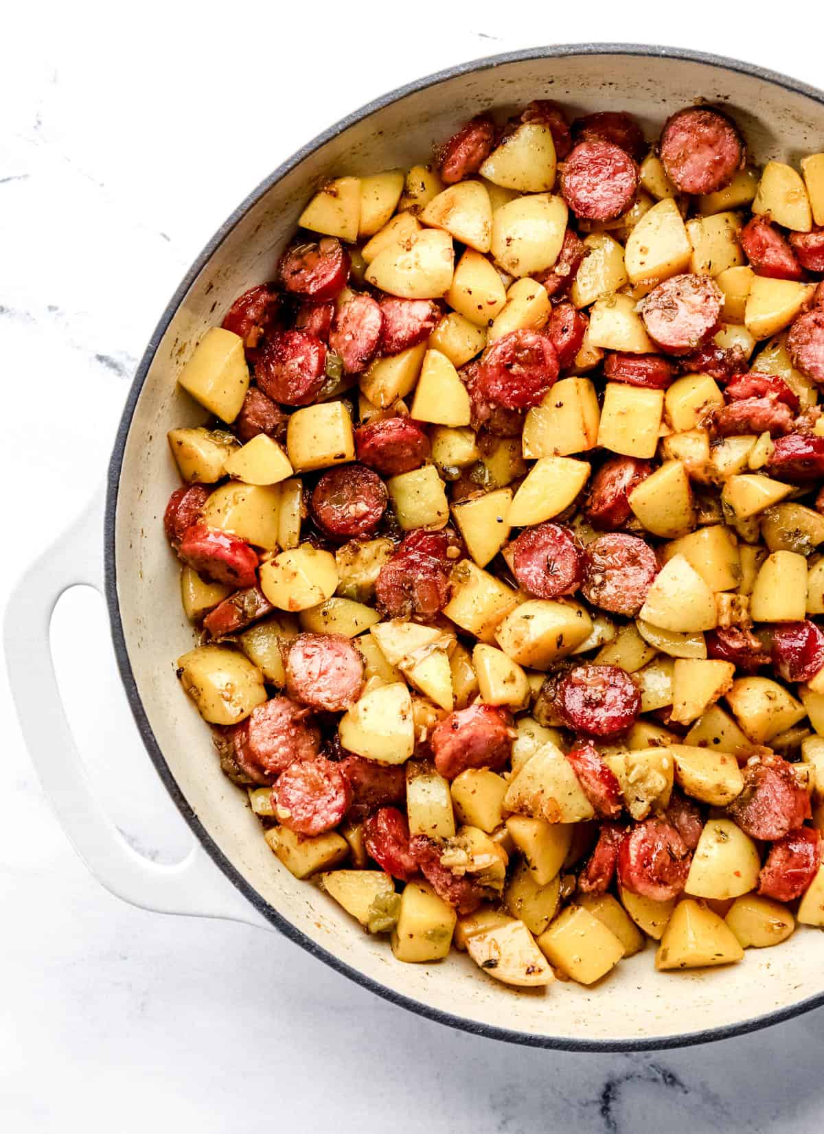 Overhead view of sausage and potatoes in white cast iron braiser. 