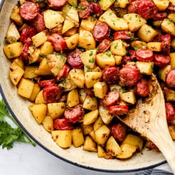 Overhead view of finished sausage and potatoes recipe in white braiser on marble surface with wooden spoon in it.