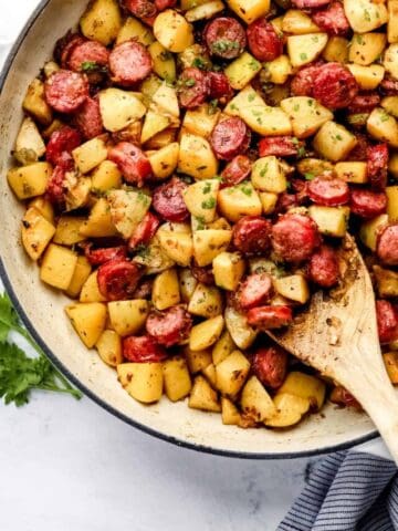 Overhead view of finished sausage and potatoes recipe in white braiser on marble surface with wooden spoon in it.