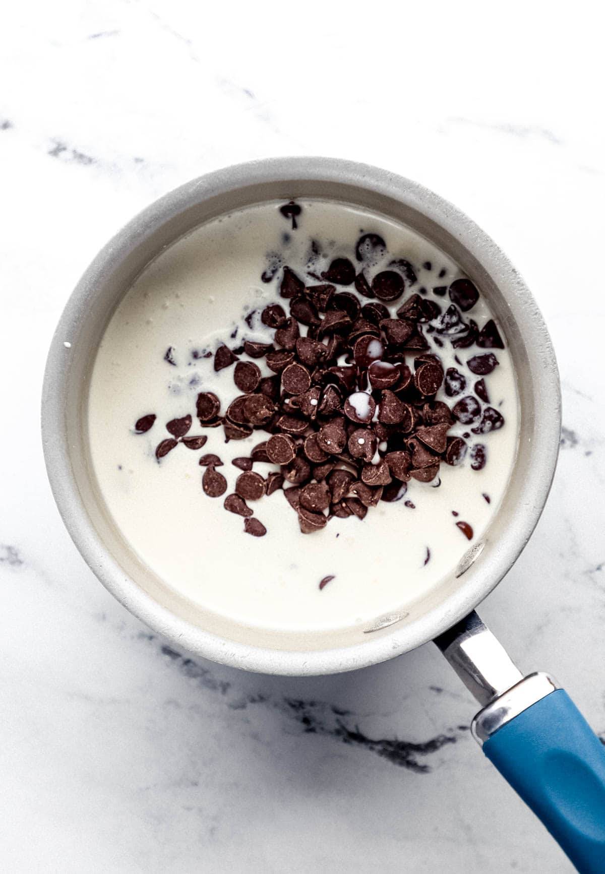 Heavy cream and chocolate chips in saucepan on marble surface.