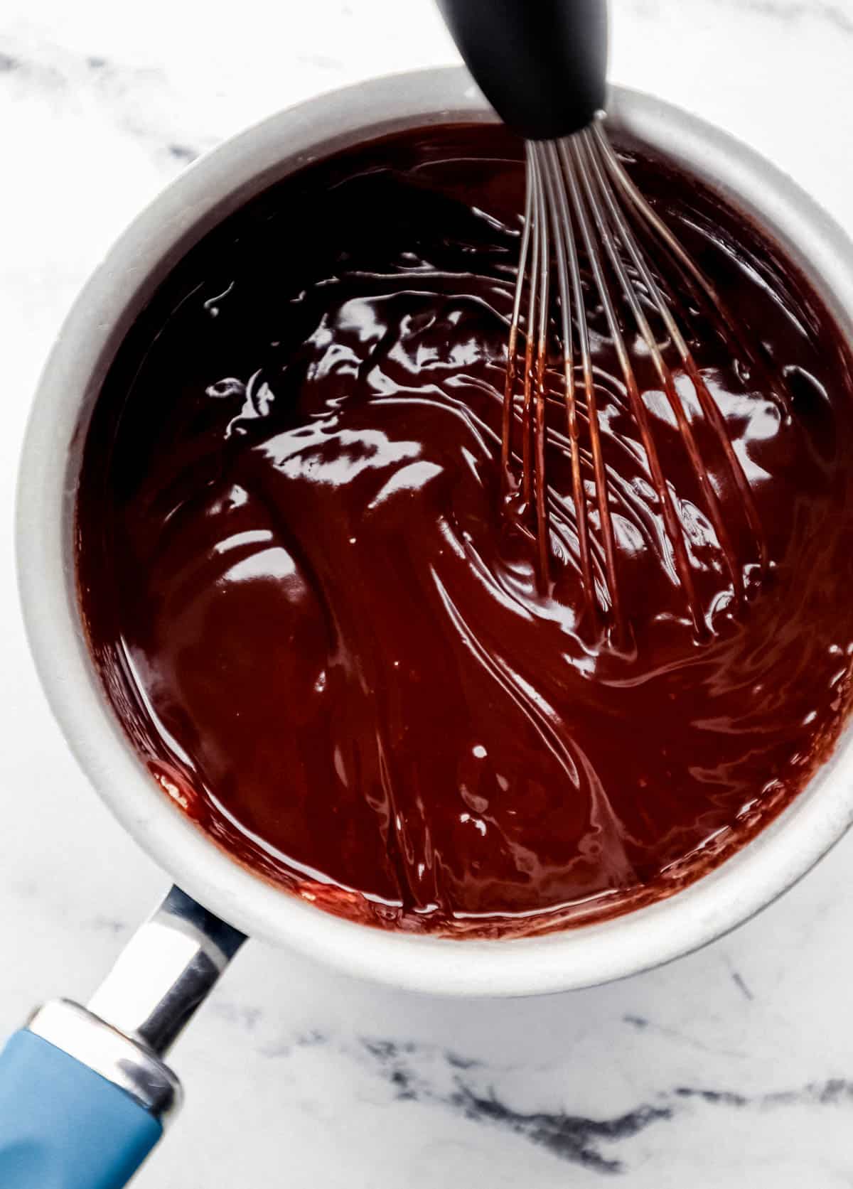 Chocolate fondue in saucepan with whisk on marble surface.