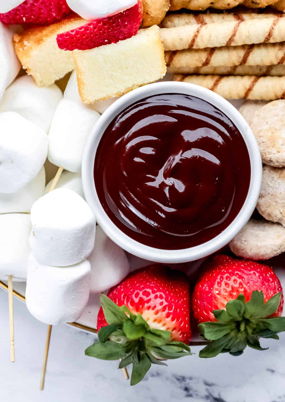 Close up view of small white bowl with chocolate fondue in it surrounded by marshmallows, cookies, and strawberries.