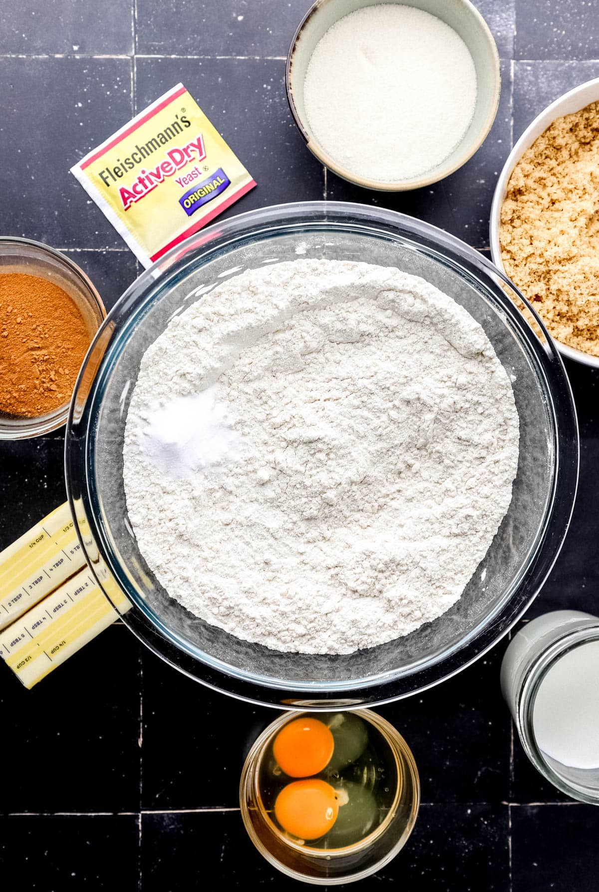 Overhead view of ingredients needed to make cinnamon rolls in separate bowls and containers on black tile surface. 
