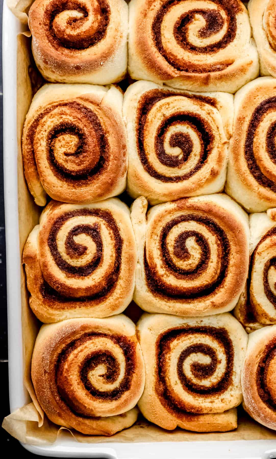 Baked cinnamon rolls in white parchment lined baking dish on black tile surface. 