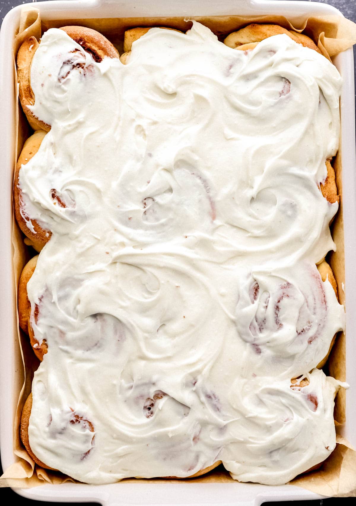 Baked cinnamon rolls topped with frosting in parchment lined baking sheet. 