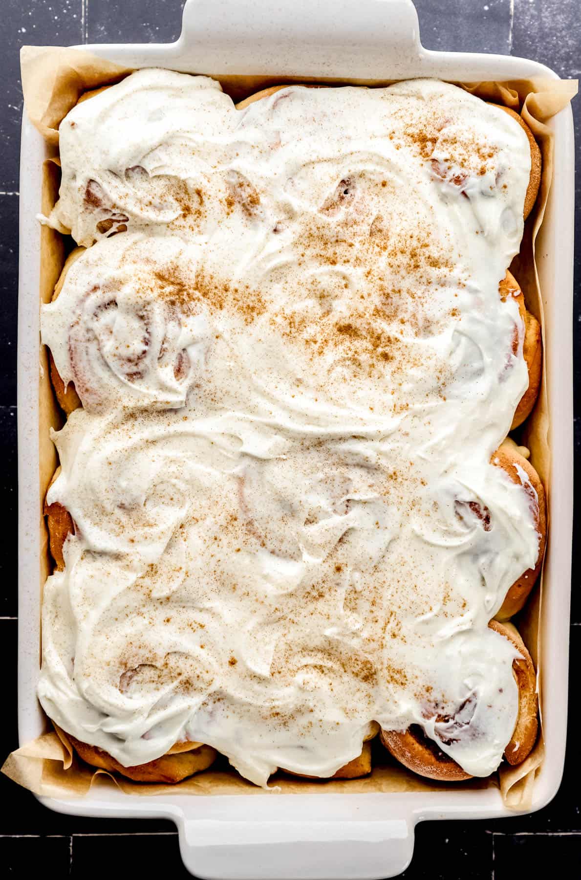 Overhead view of finished cinnamon rolls in rectangle parchment lined baking dish, topped with frosting and cinnamon. 