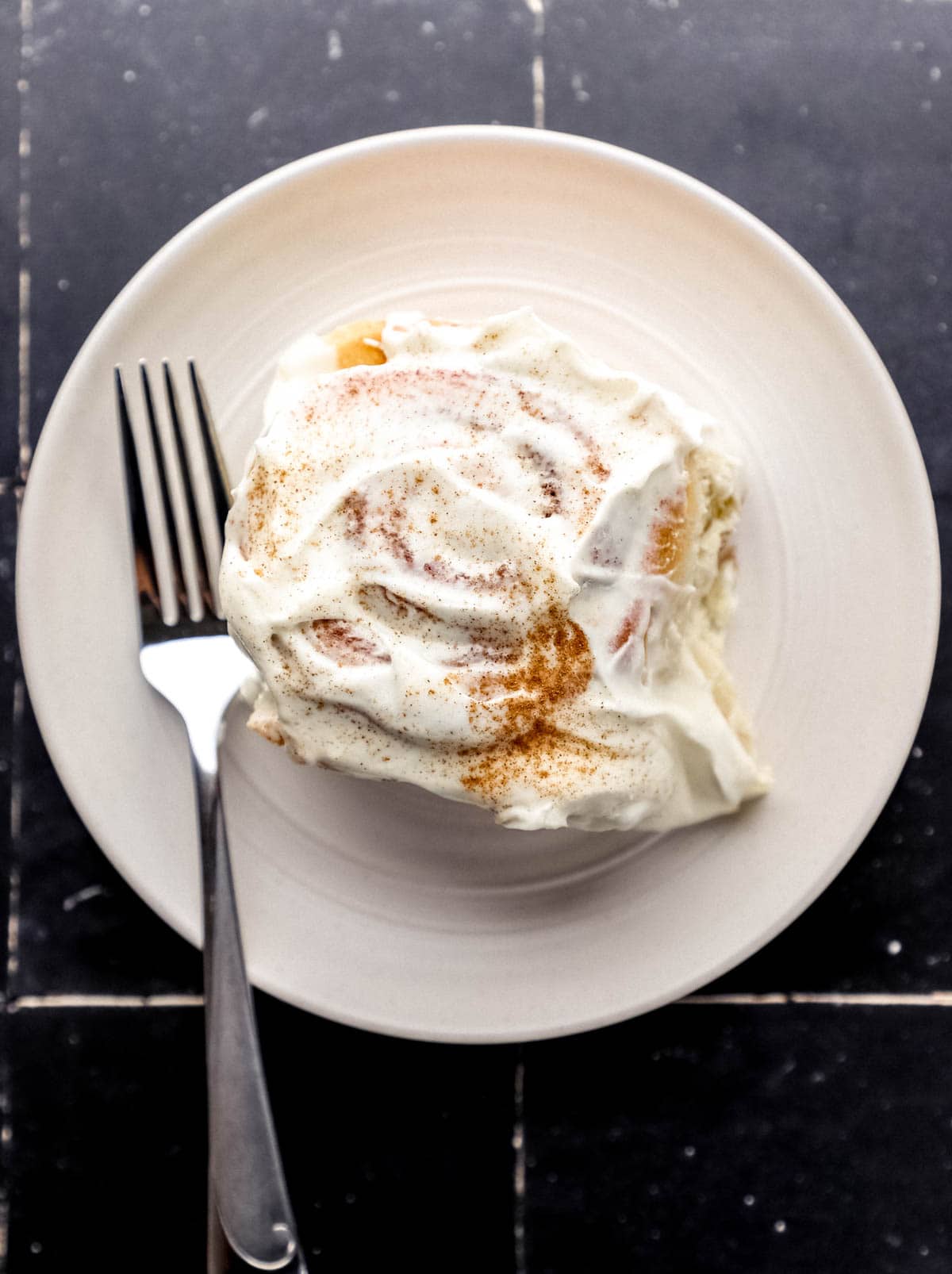 Small white plate with single cinnamon roll and fork on it on black tile surface. 