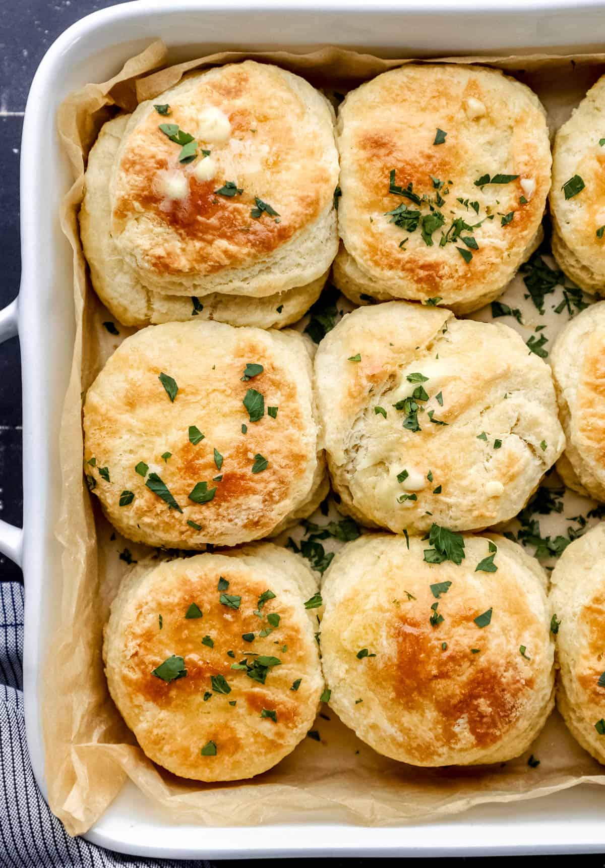 Overhead view of biscuits in parchment lined baking dish topped with butter and chopped parsley. 