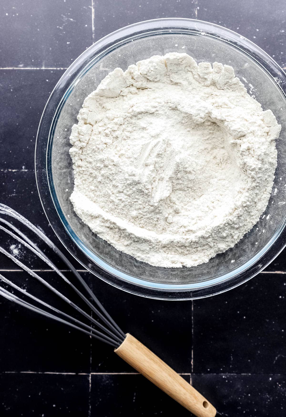 Overhead view of large glass bowl with dry ingredients in it by a whisk. 