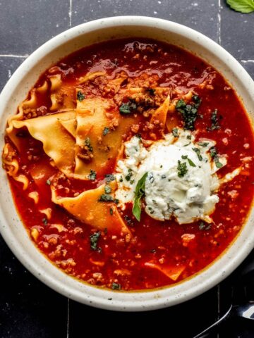 Overhead view of bowl of lasagna soup on black tile marble surface by a spoon and cloth napkin.