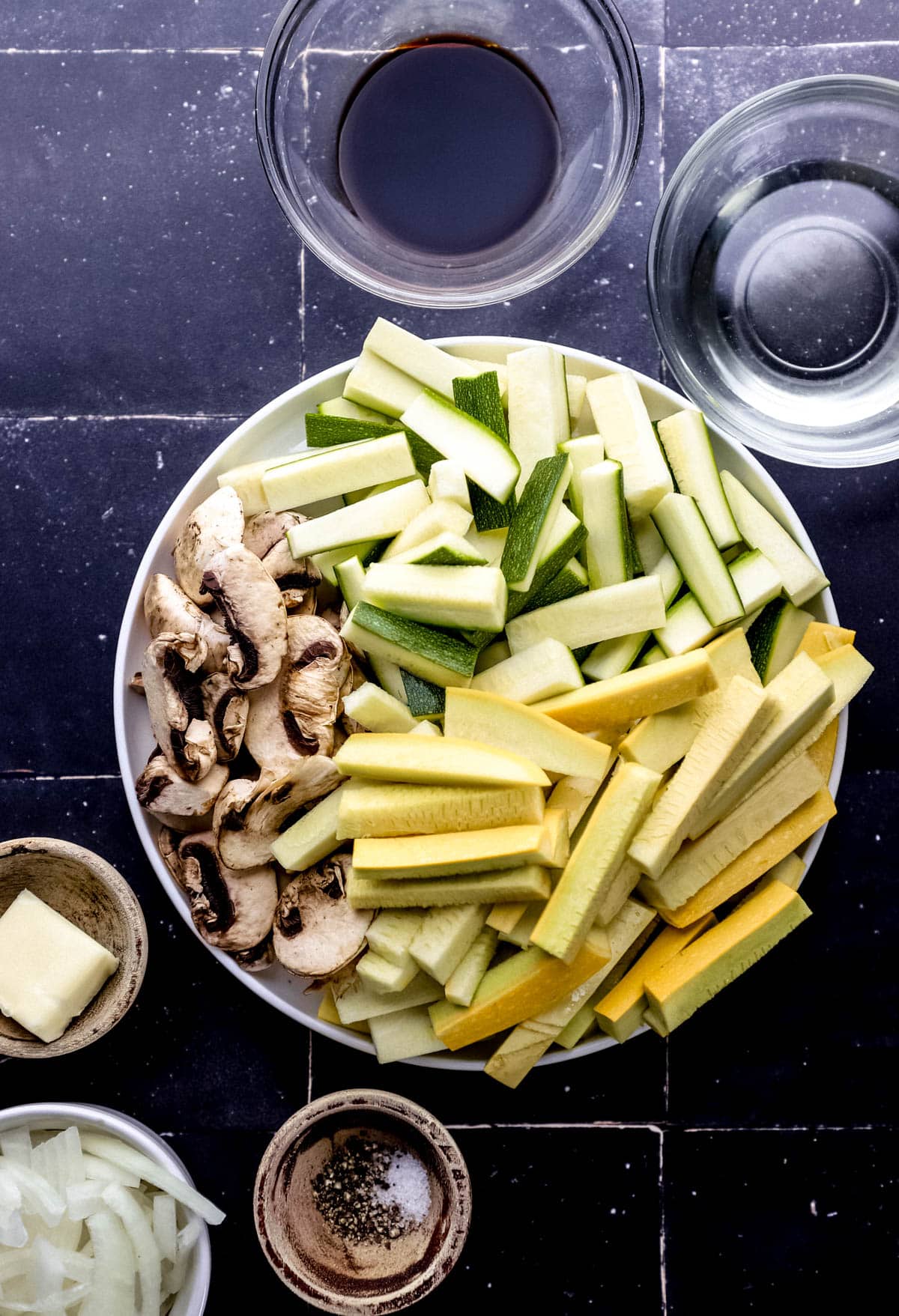 Overhead view of ingredients needed to make vegetables in separate bowls on black tile surface. 