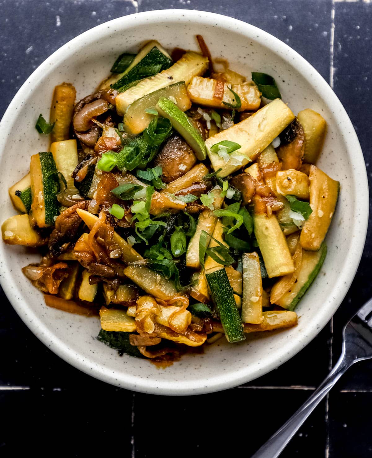 Overhead view of white bowl of hibachi vegetables topped with chopped green onion on black tile surface. 