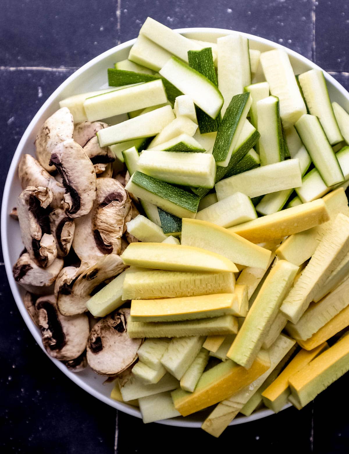 Large white plate with fresh sliced zucchini, squash, and sliced mushrooms on it. 