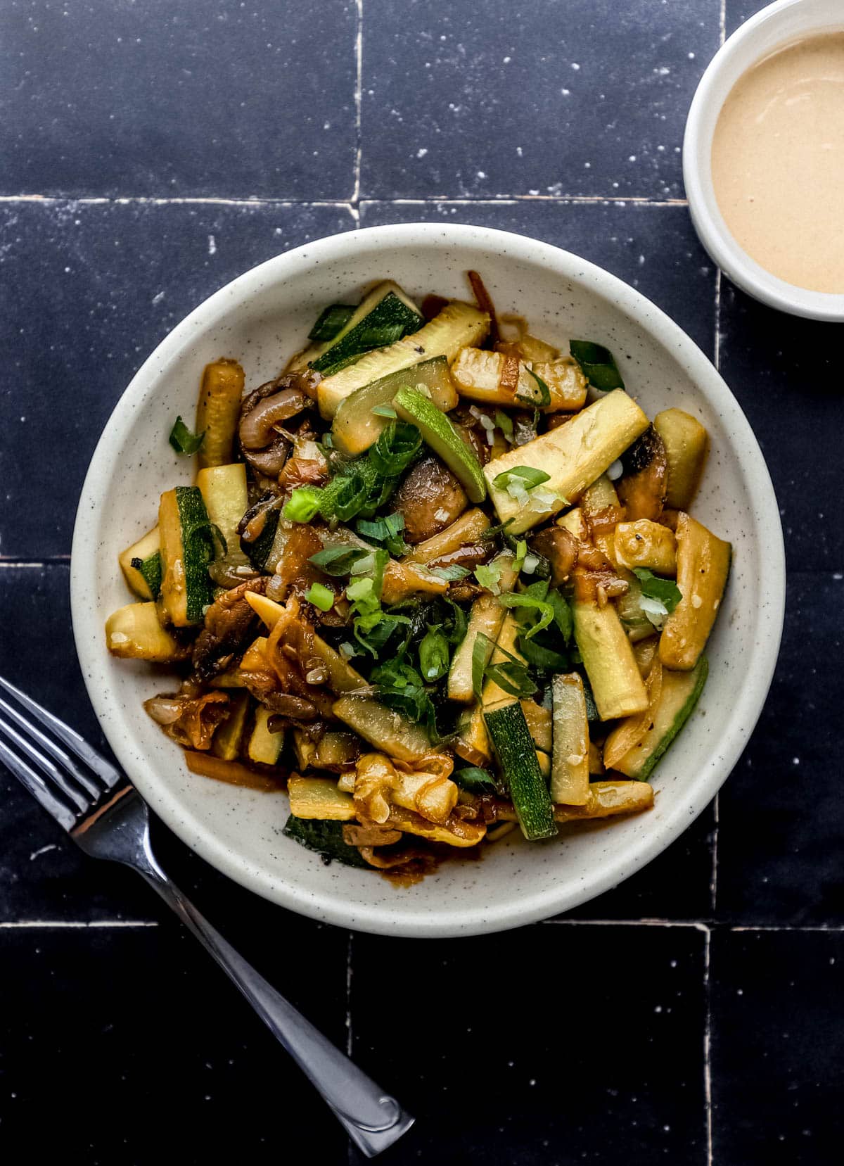 Overhead view of bowl of hibachi vegetables on black tile surface beside a fork and small bowl of sauce. 