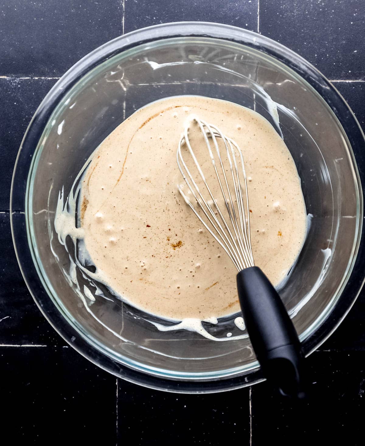 Overhead view of sauce ingredients combined in large glass bowl with whisk on black tile surface. 