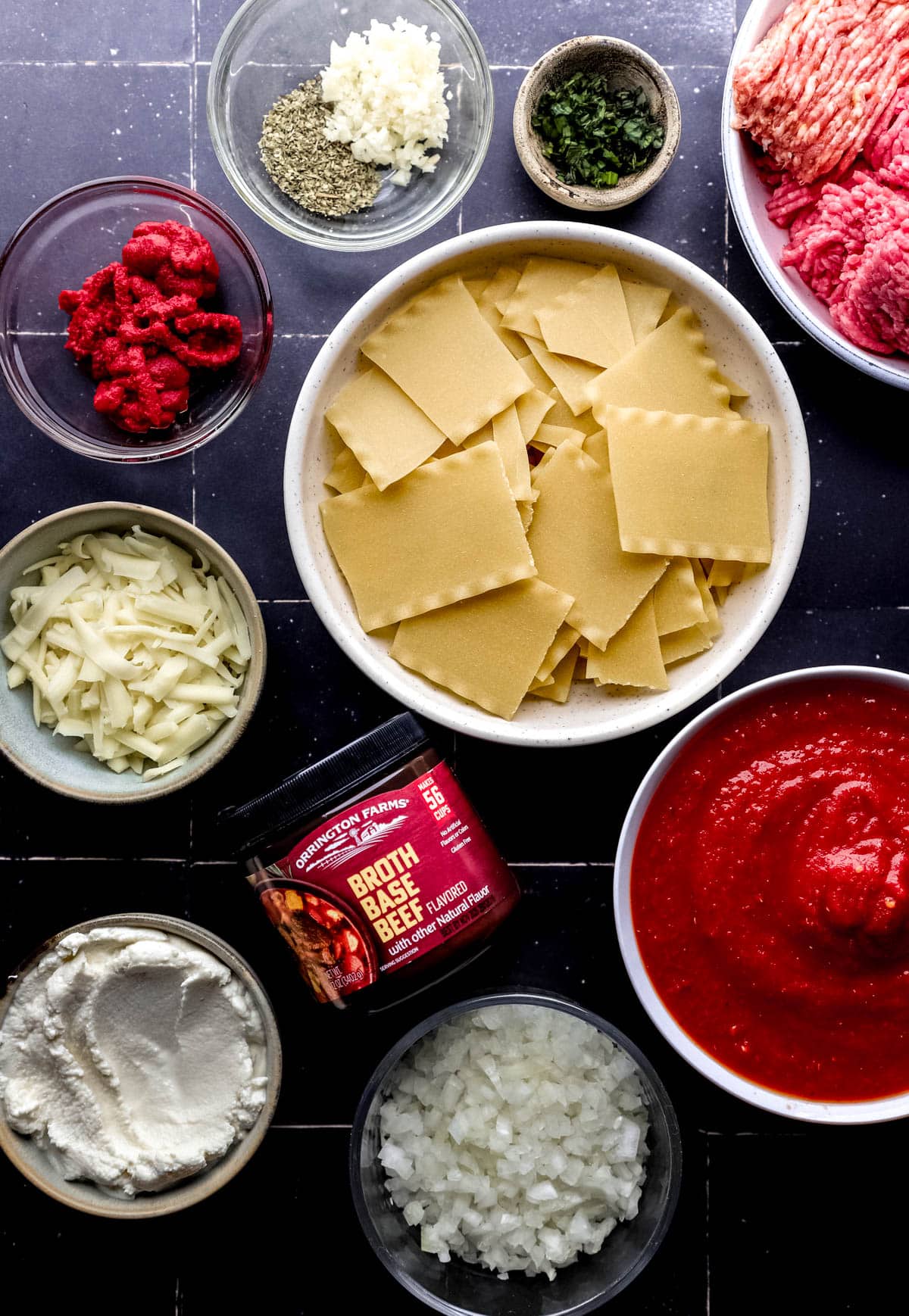 Overhead view of ingredients needed to make lasagna soup in separate bowls and containers on black tile surface. 