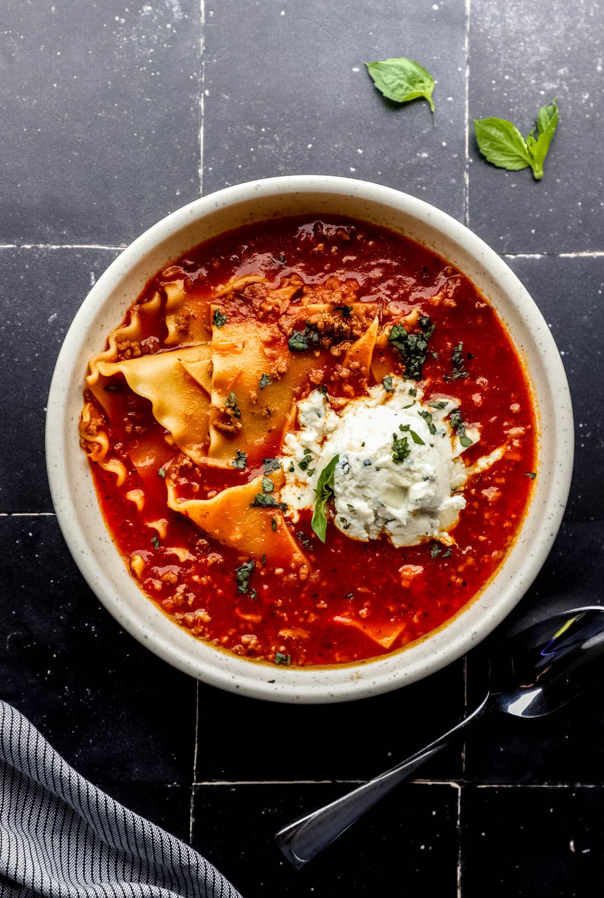 Overhead view of bowl of soup beside a spoon and cloth napkin on black tile surface. 