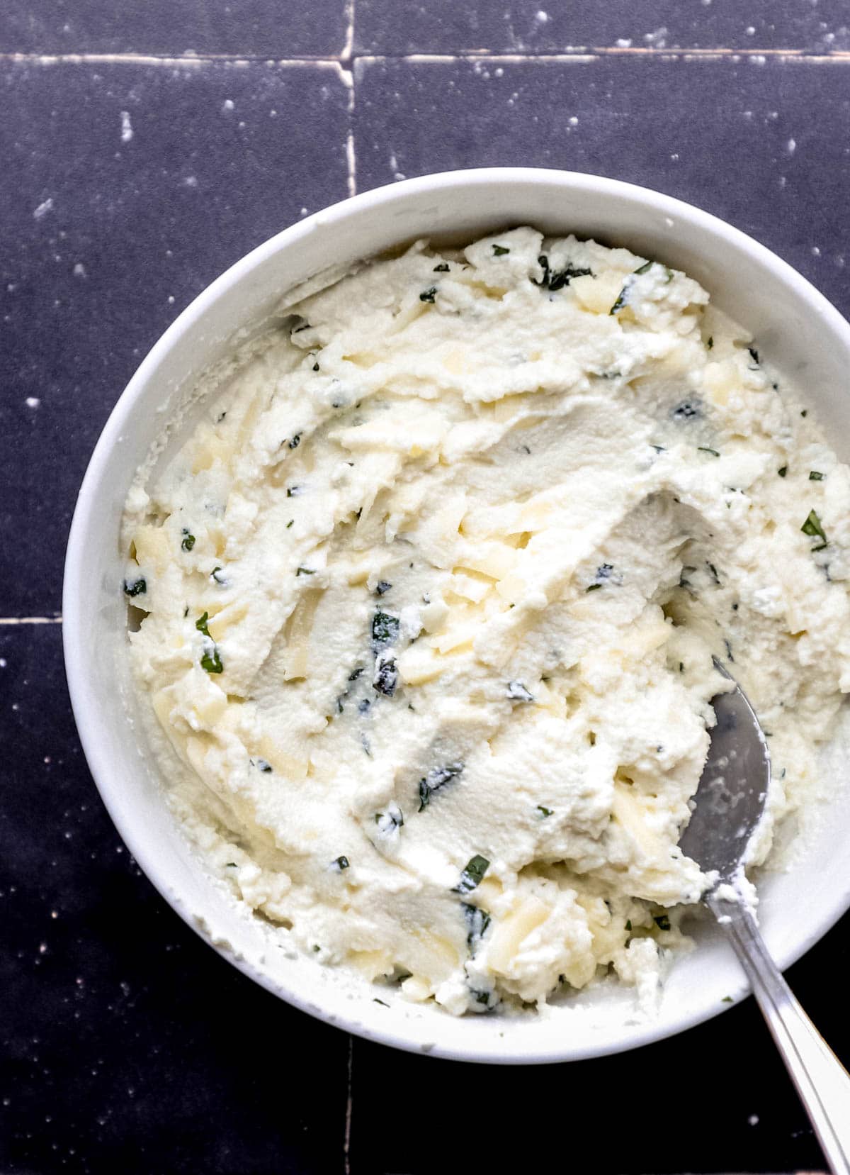 Overhead view of cheese mixture in white bowl with spoon on black tile surface. 